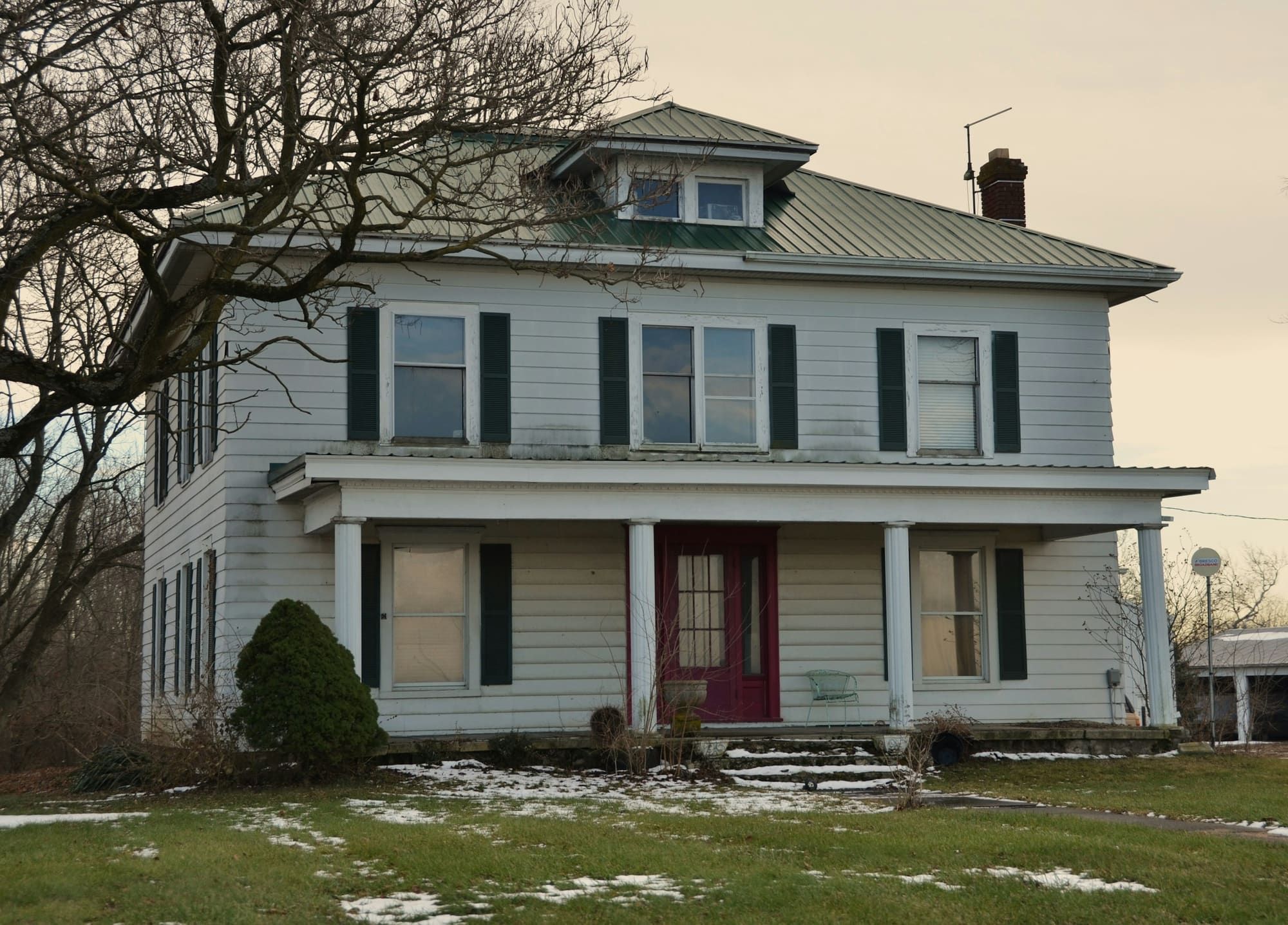 White two-story farmhouse with a metal hip roof and patches of snow on the lawn