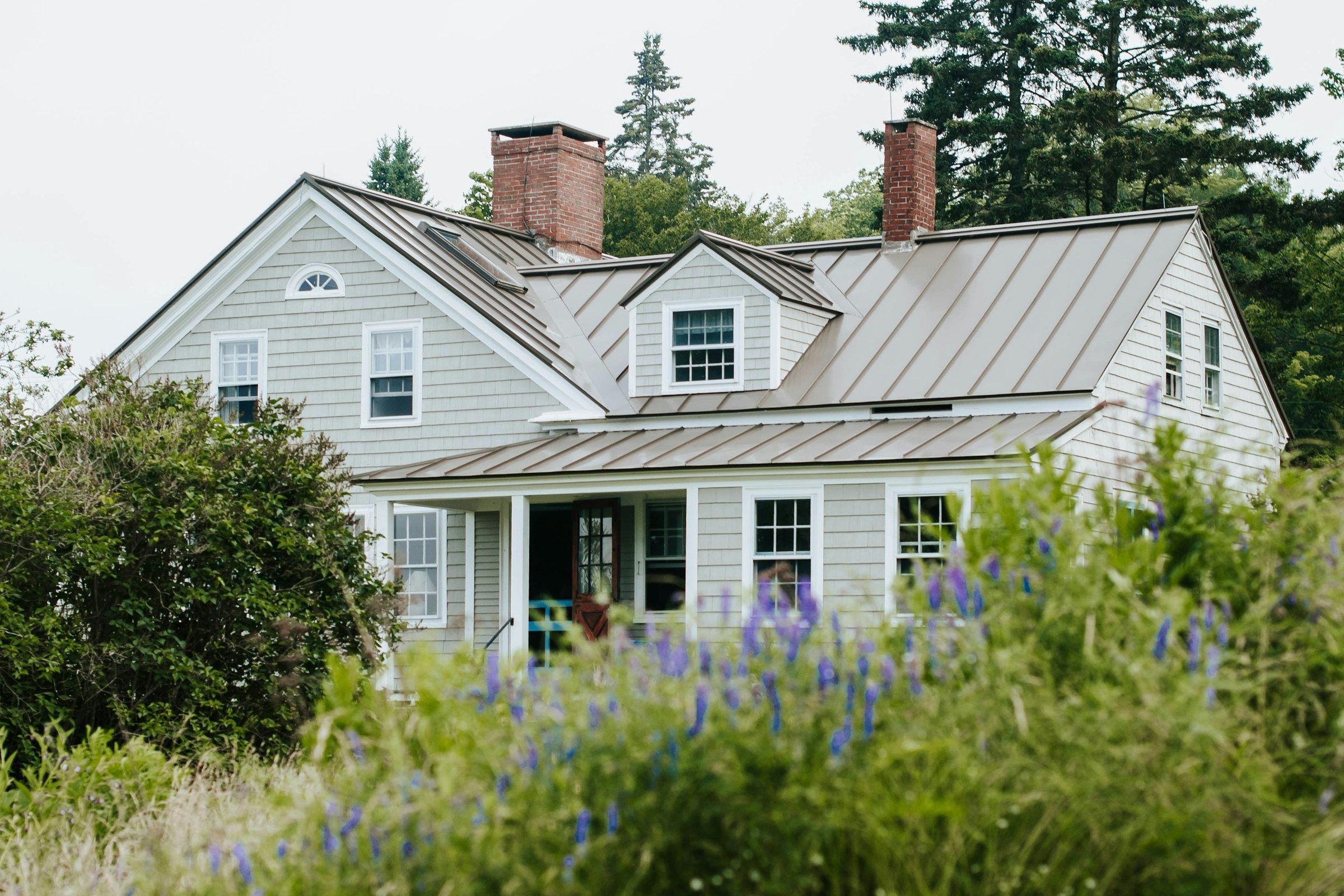 Gray clapboard farmhouse with a standing seam metal roof and brick chimneys