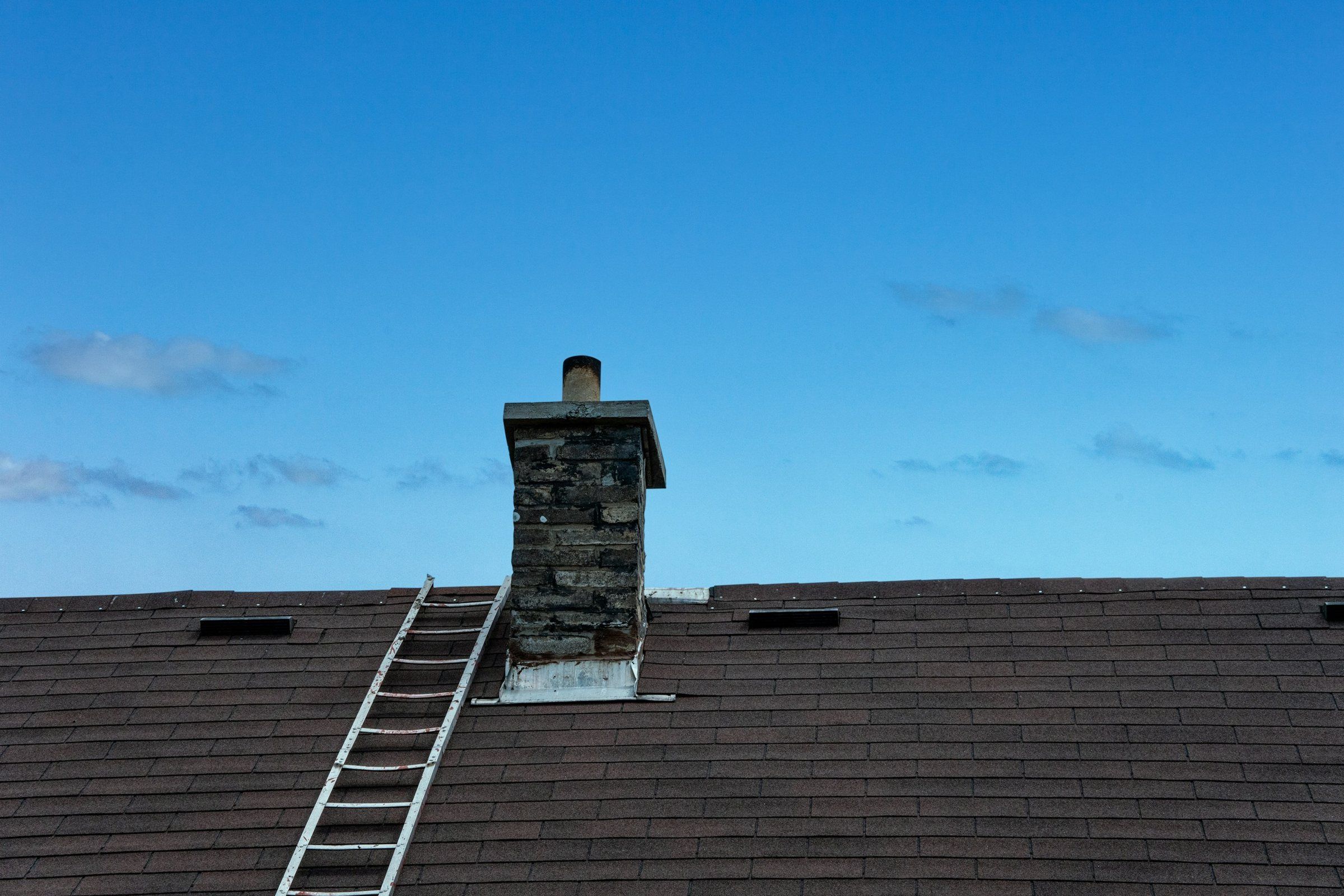 Ladder on an asphalt shingle roof next to a stone chimney with metal flashing at its base