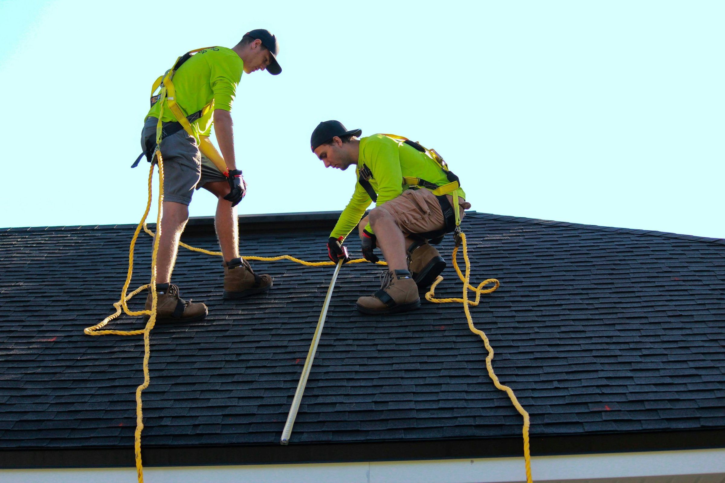 Two roofers in safety harnesses working on a dark asphalt shingle roof