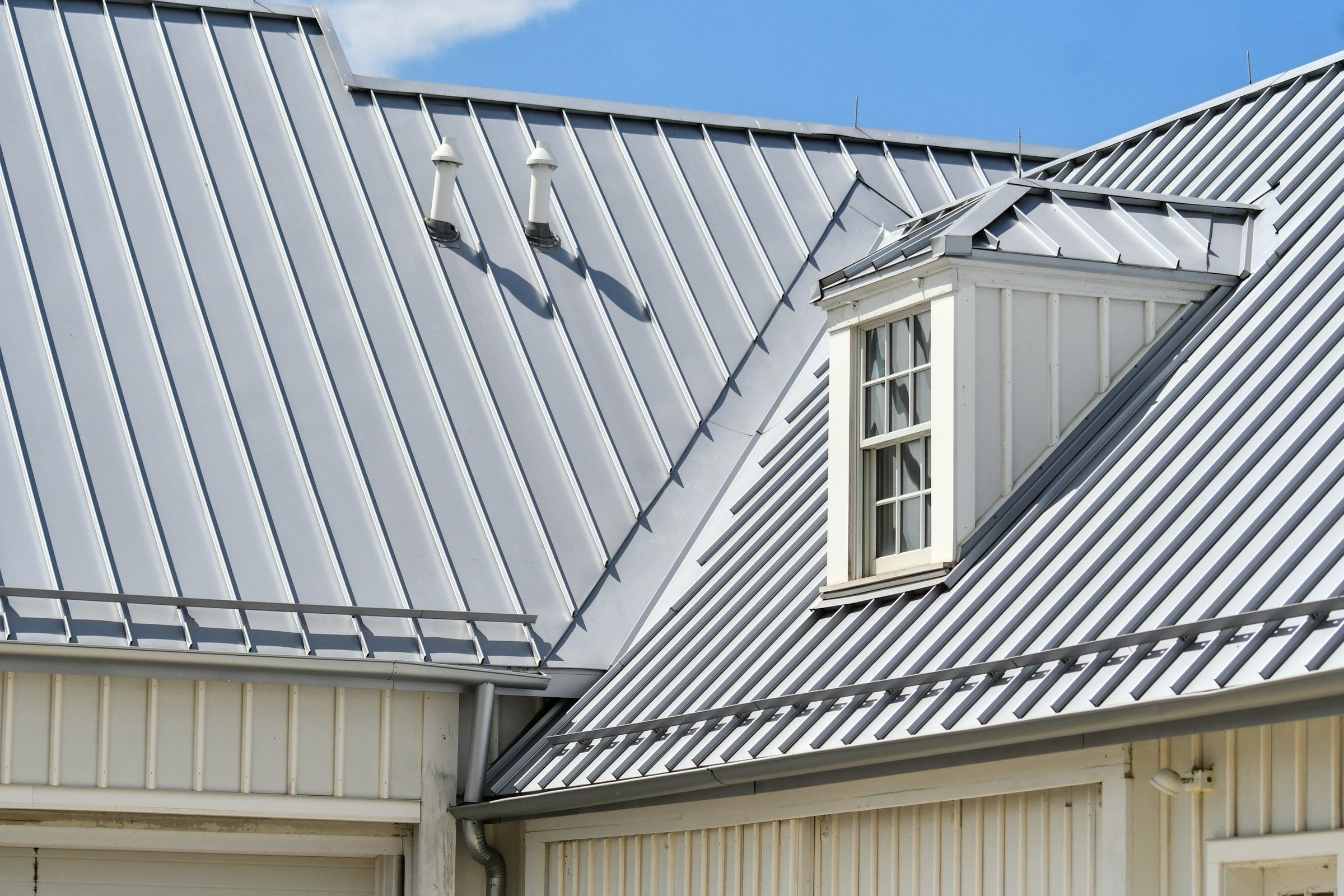 Gray standing seam metal roof with a dormer window, vent pipes and snow guard rails above the eaves