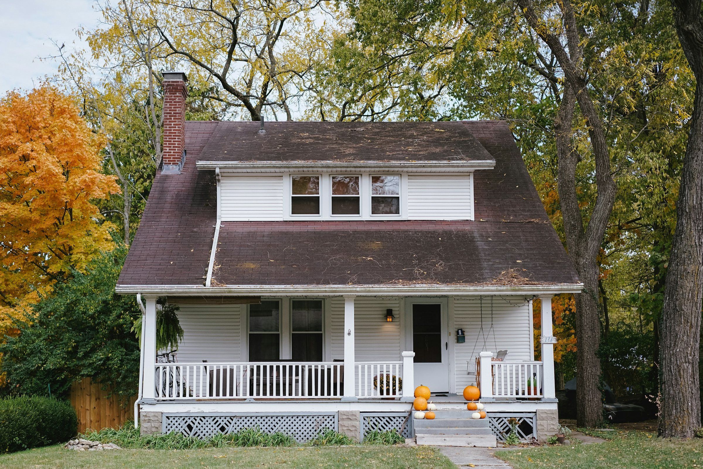 White bungalow with a front porch and an aging shingle roof scattered with leaf debris